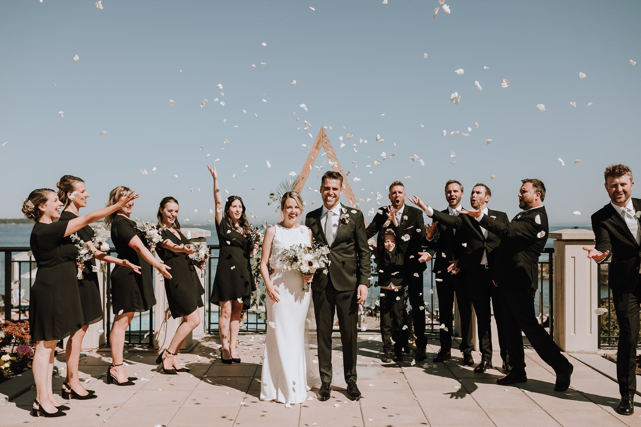 A group of people outdoors, some holding bouquets, with a clear sky and a body of water in the background.
