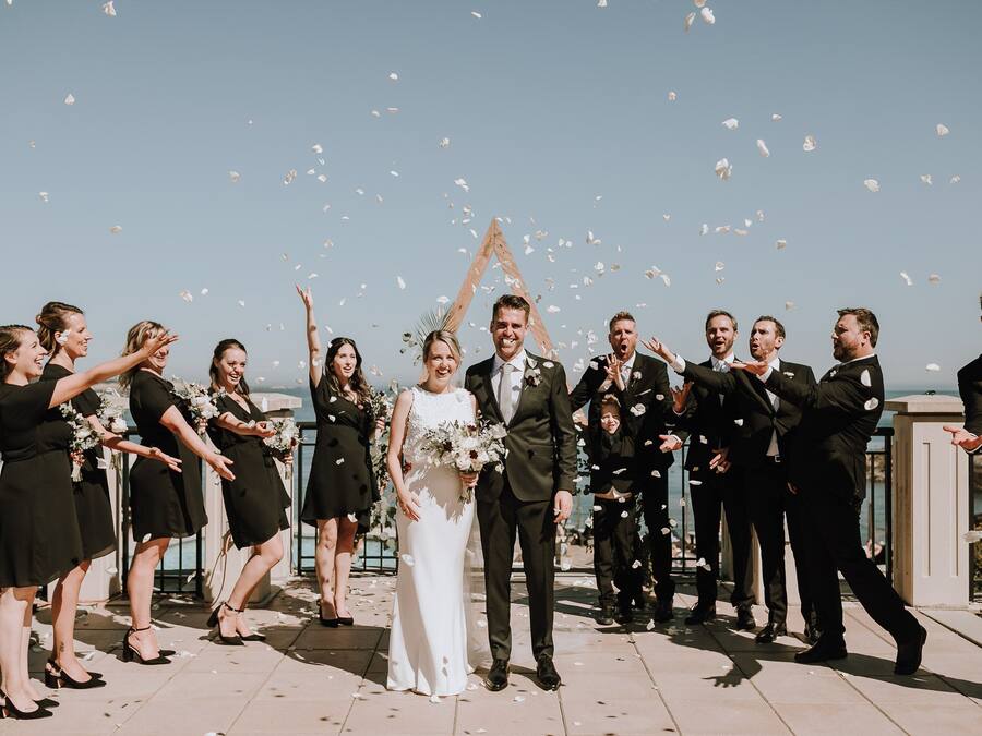 A group of people outdoors, some holding bouquets, with a clear sky and a body of water in the background.