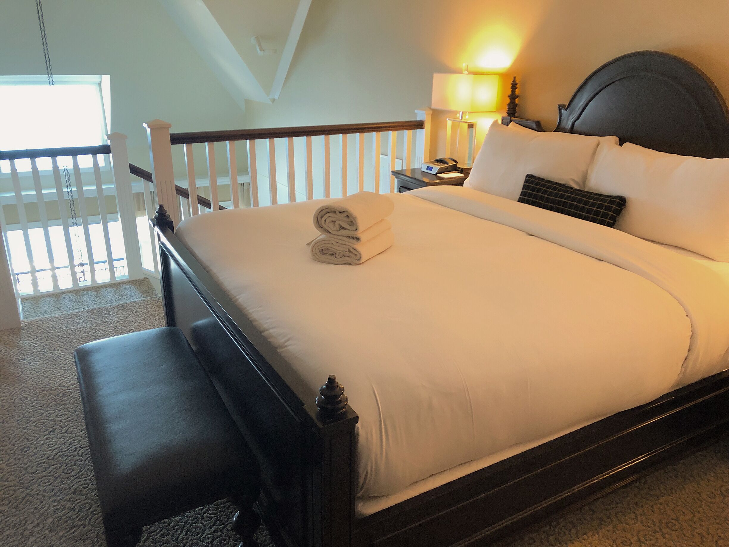 A neatly made bed with a pillow and a rolled towel, a bedside table with a lamp, and a view of a balcony with white railings.