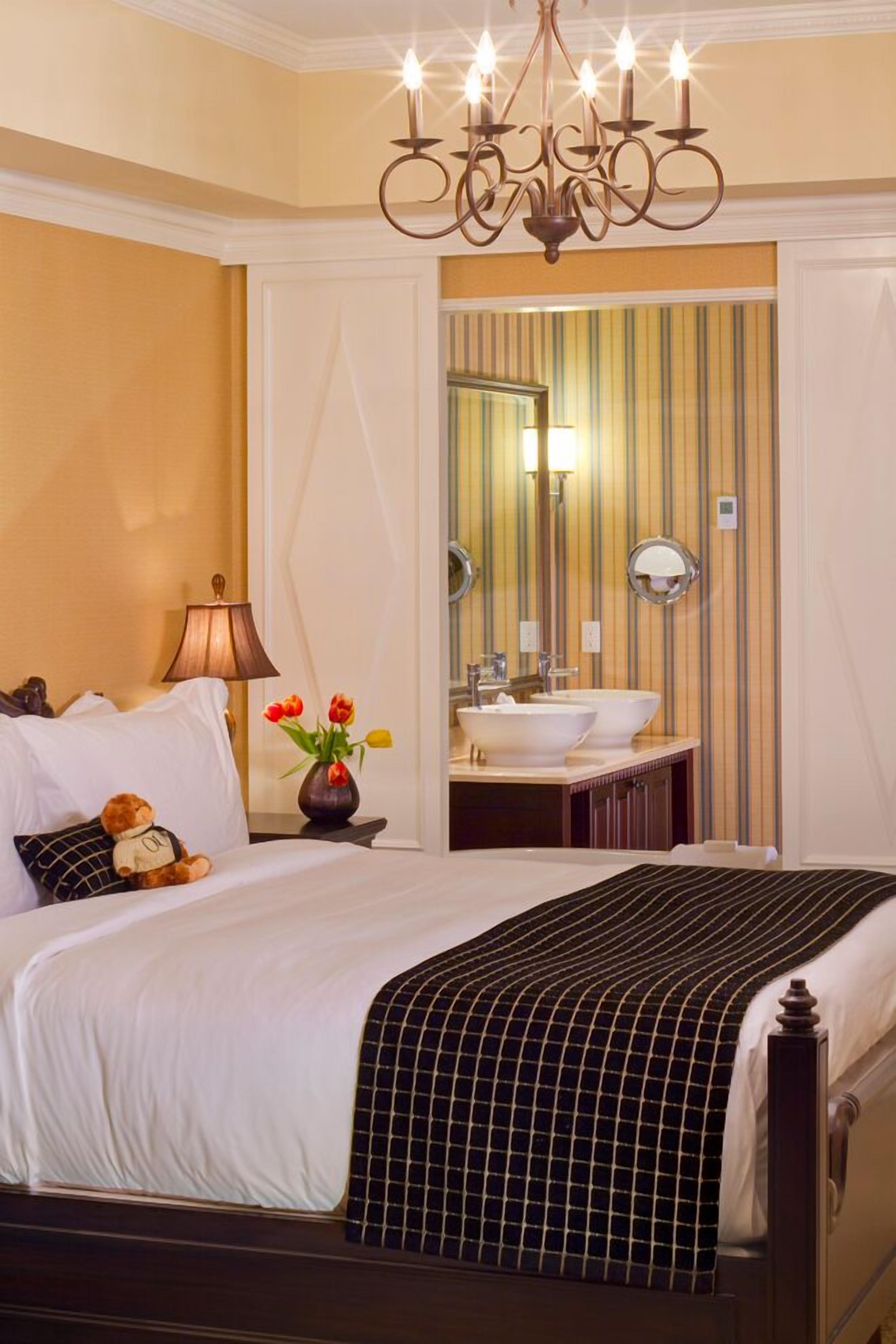 A hotel room with a bed, a bedside lamp, a teddy bear, a vase with flowers, a chandelier, a mirror, and a bathroom with a sink and mirror.