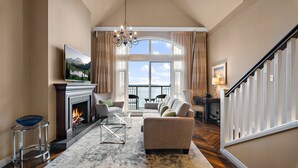 A spacious living room with a fireplace, a large window with a view of the water, a chandelier, and a staircase. - Oak Bay Beach Hotel (Victoria)