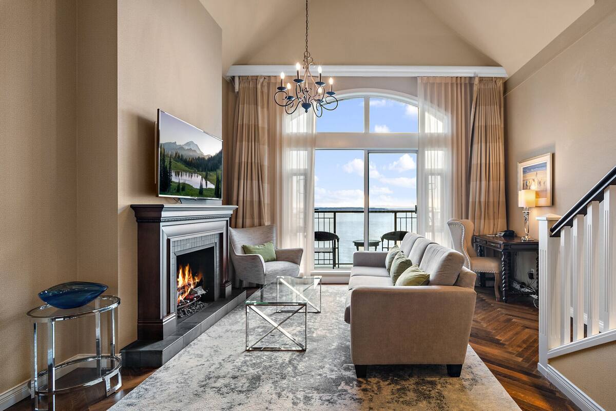 Ein geräumiges Wohnzimmer mit Kamin, einem großen Fenster mit Blick auf das Wasser, einer Kristallleuchtre und einer Treppe.
