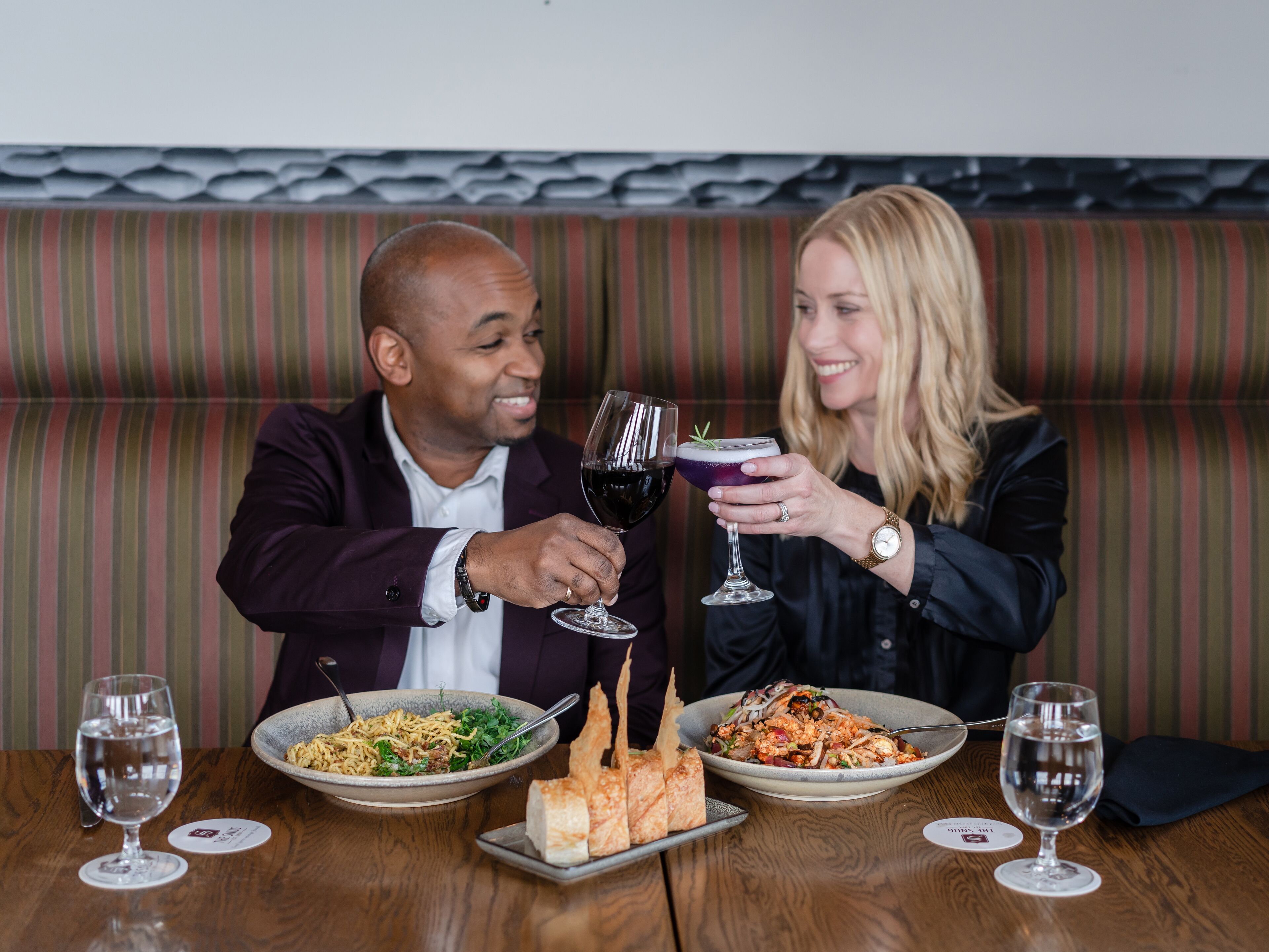 Un homme et une femme sont assis à une table en bois devant deux assiettes de nourriture, deux verres de vin et un verre à cocktail.