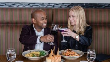 Un homme et une femme sont assis à une table en bois devant deux assiettes de nourriture, deux verres de vin et un verre à cocktail.