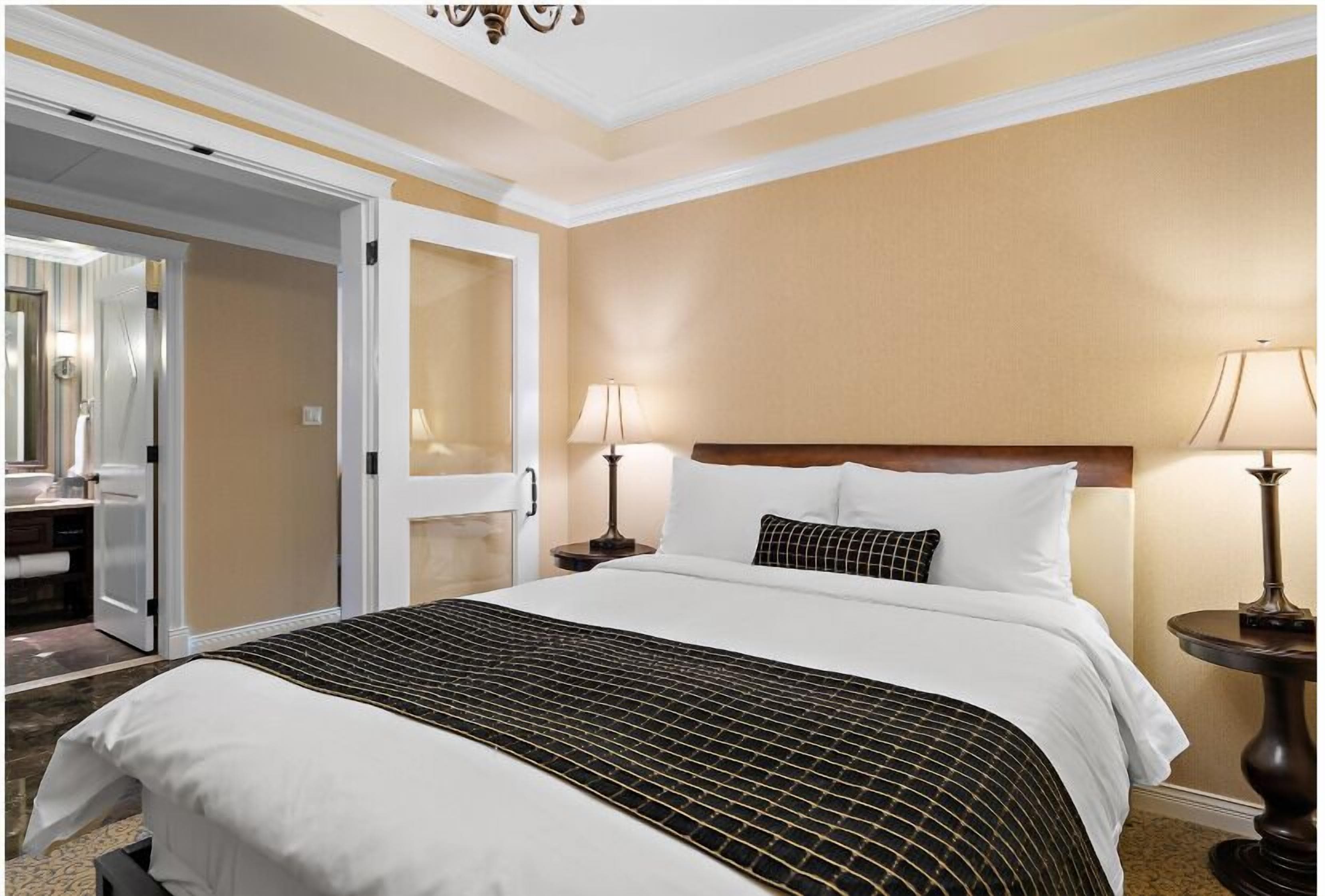 A hotel room with a bed, two bedside lamps, a nightstand, a mirror, and a door leading to a bathroom.