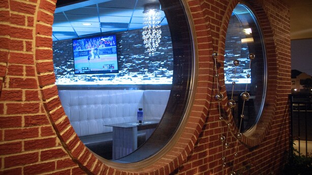 A brick-walled room with a large circular window reflecting a television screen, a white cushioned bench, and a hanging decorative light fixture. - Unique Suites Hotel (Charleston)