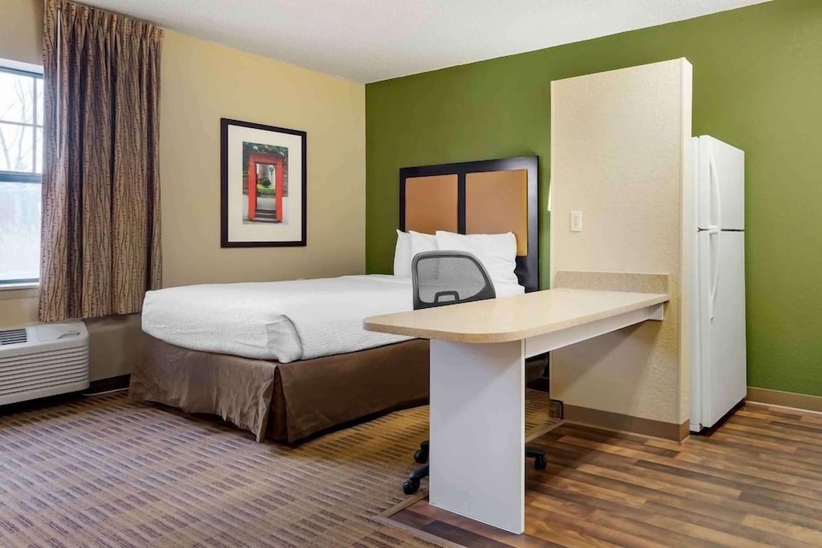 A hotel room with a bed, a desk, a chair, a refrigerator, and a window with curtains.