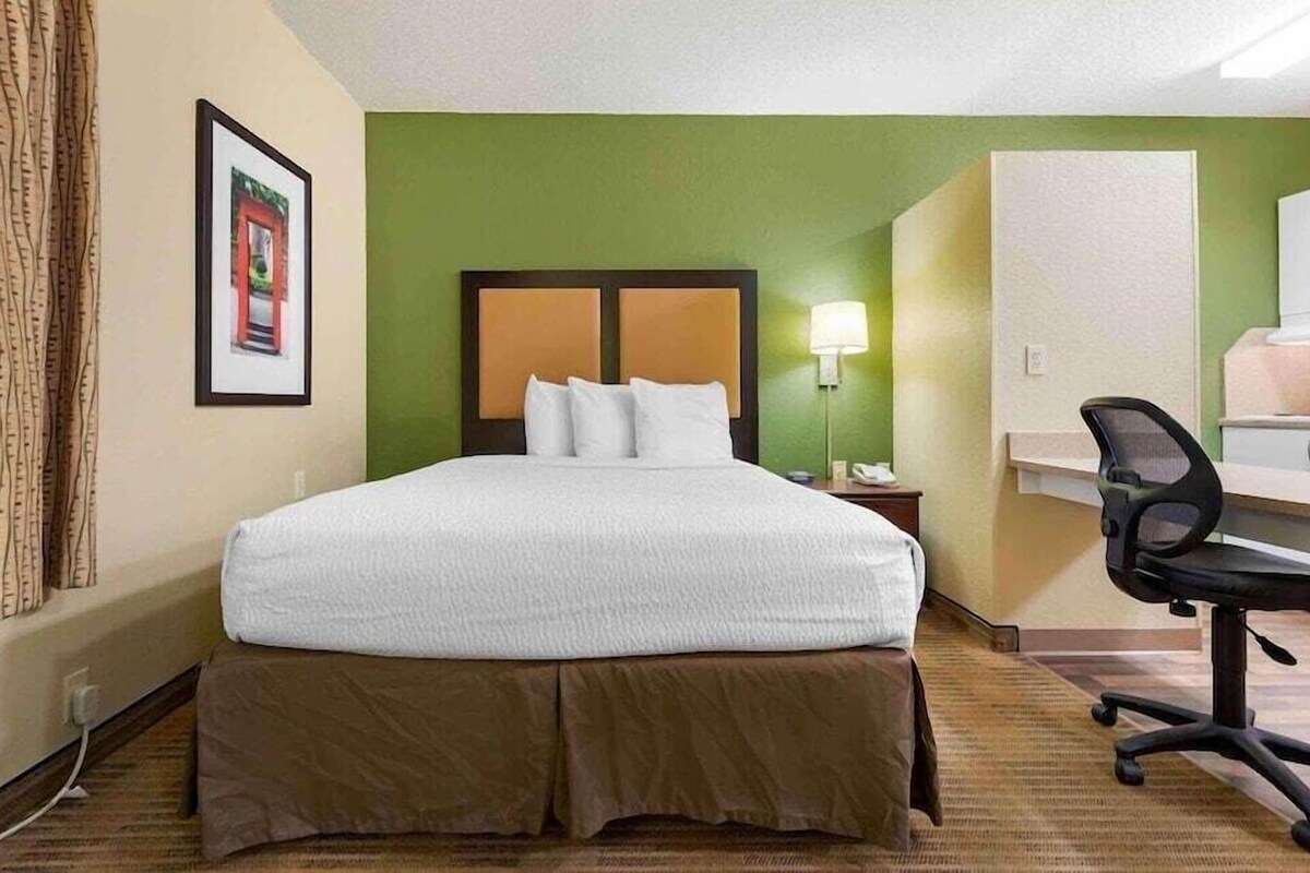 A hotel room with a bed, a desk, a chair, a nightstand, and a lamp.