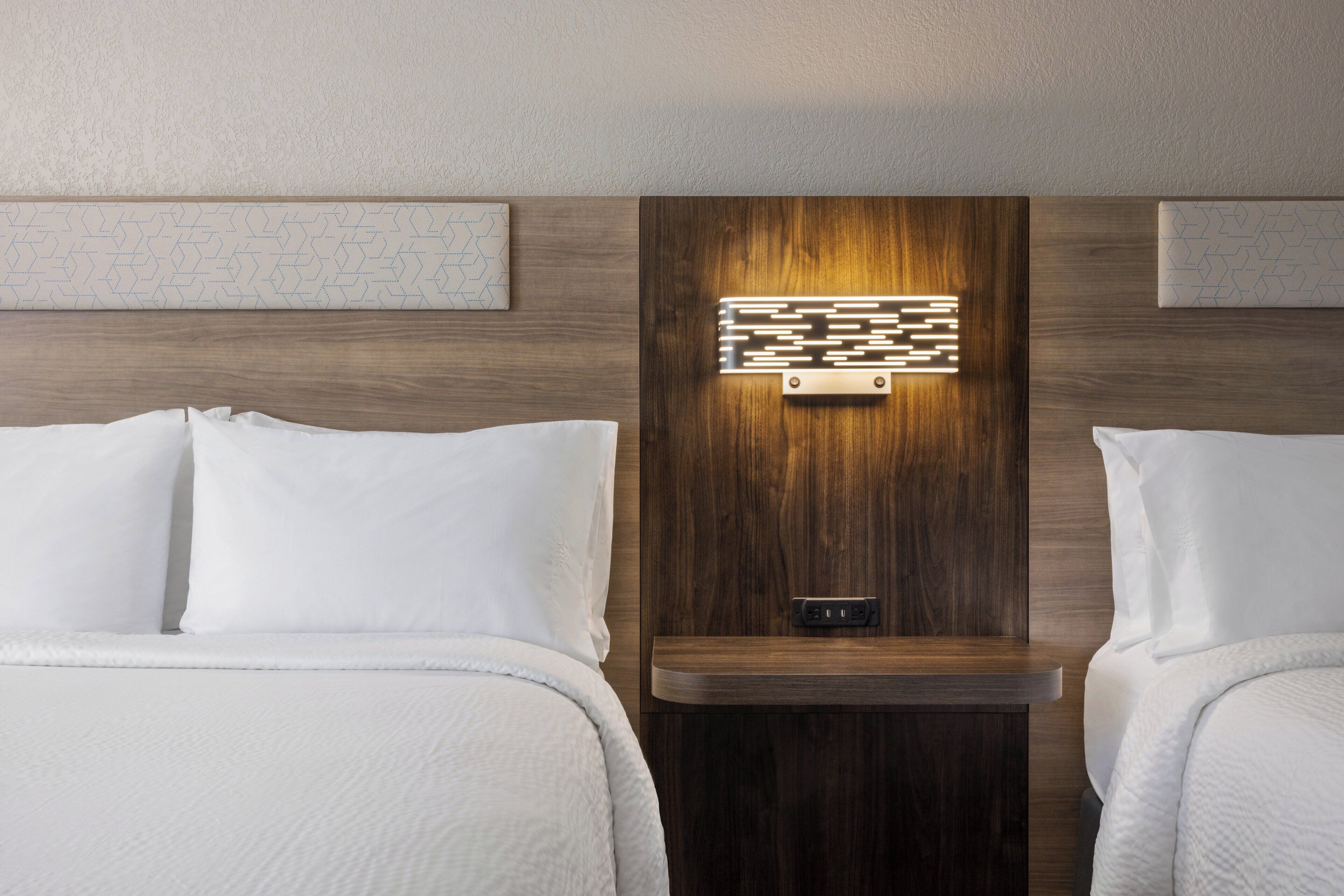 Habitación de hotel con dos camas, cabecero de madera con panel de luz y mesita de noche pequeña con toma de corriente.