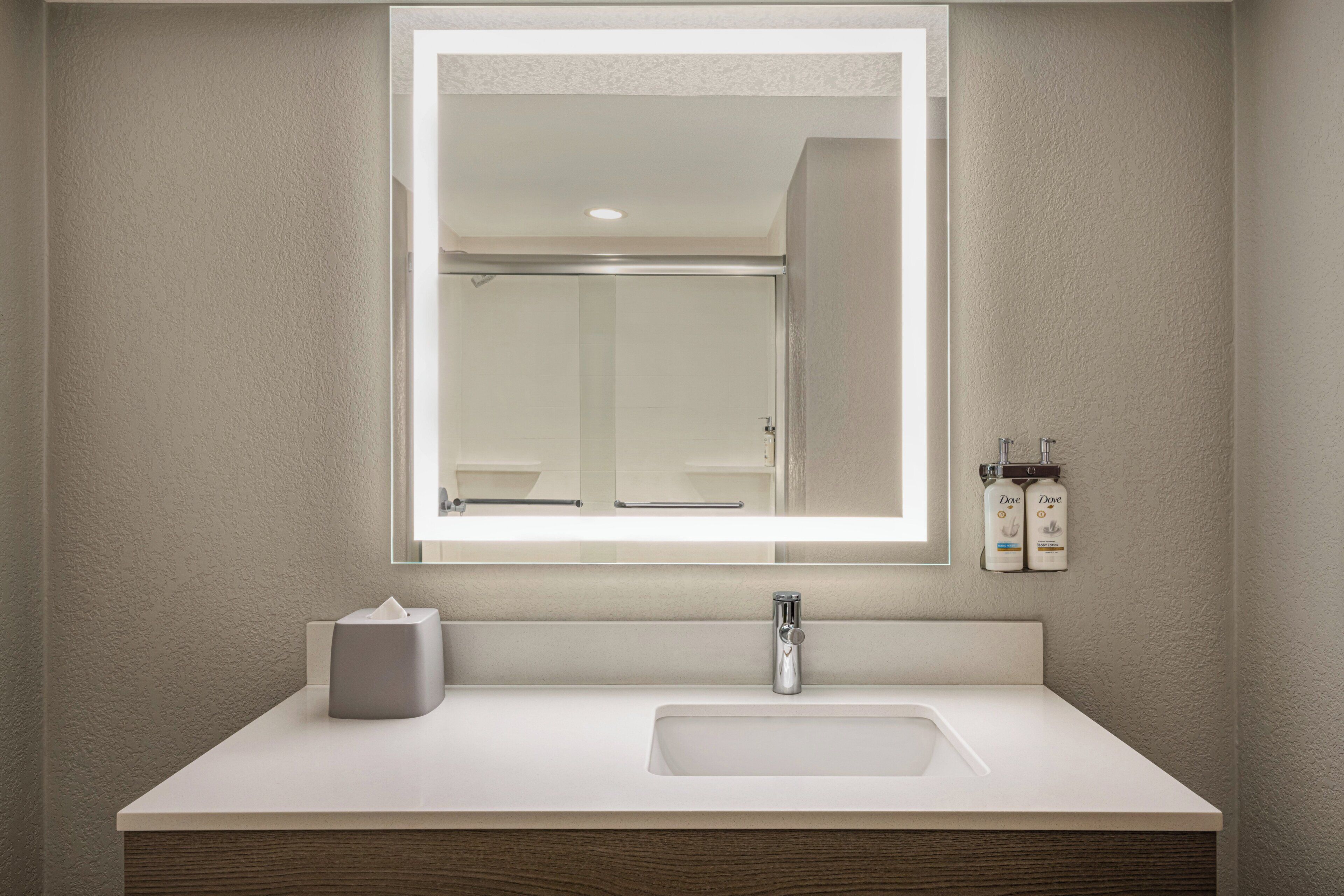 A bathroom with a large mirror, a sink, a towel dispenser, and a bottle of Dove soap.