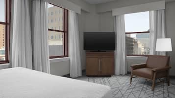 A hotel room with a large bed, a television on a wooden cabinet, a chair, and a view of the city through the windows.