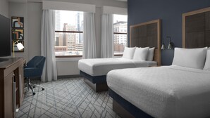 A hotel room with two beds, a desk with a chair, a TV, and large windows with curtains.