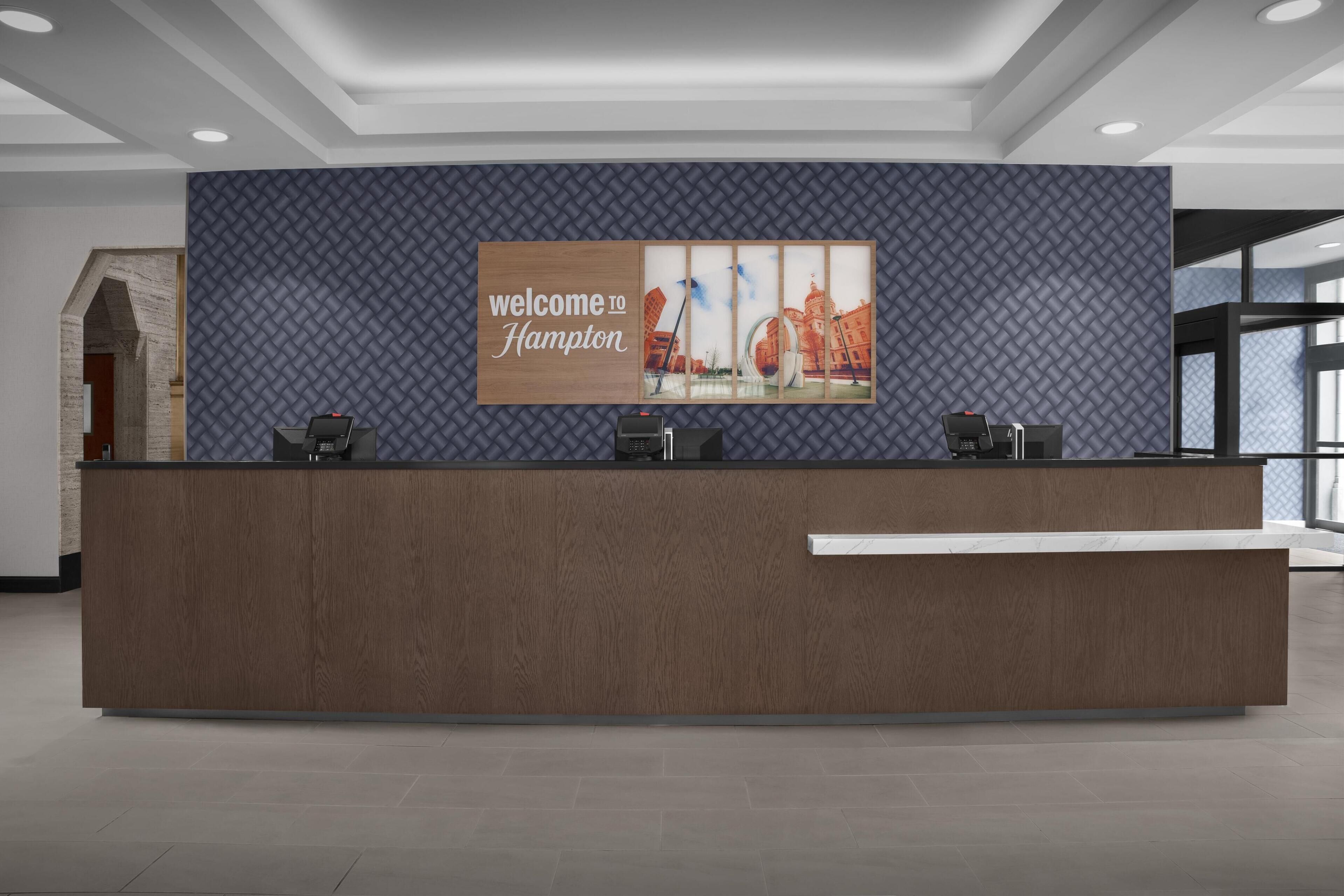 A reception desk with a "Welcome to Hampton" sign, a modern design, and a view of the cityscape through large windows.