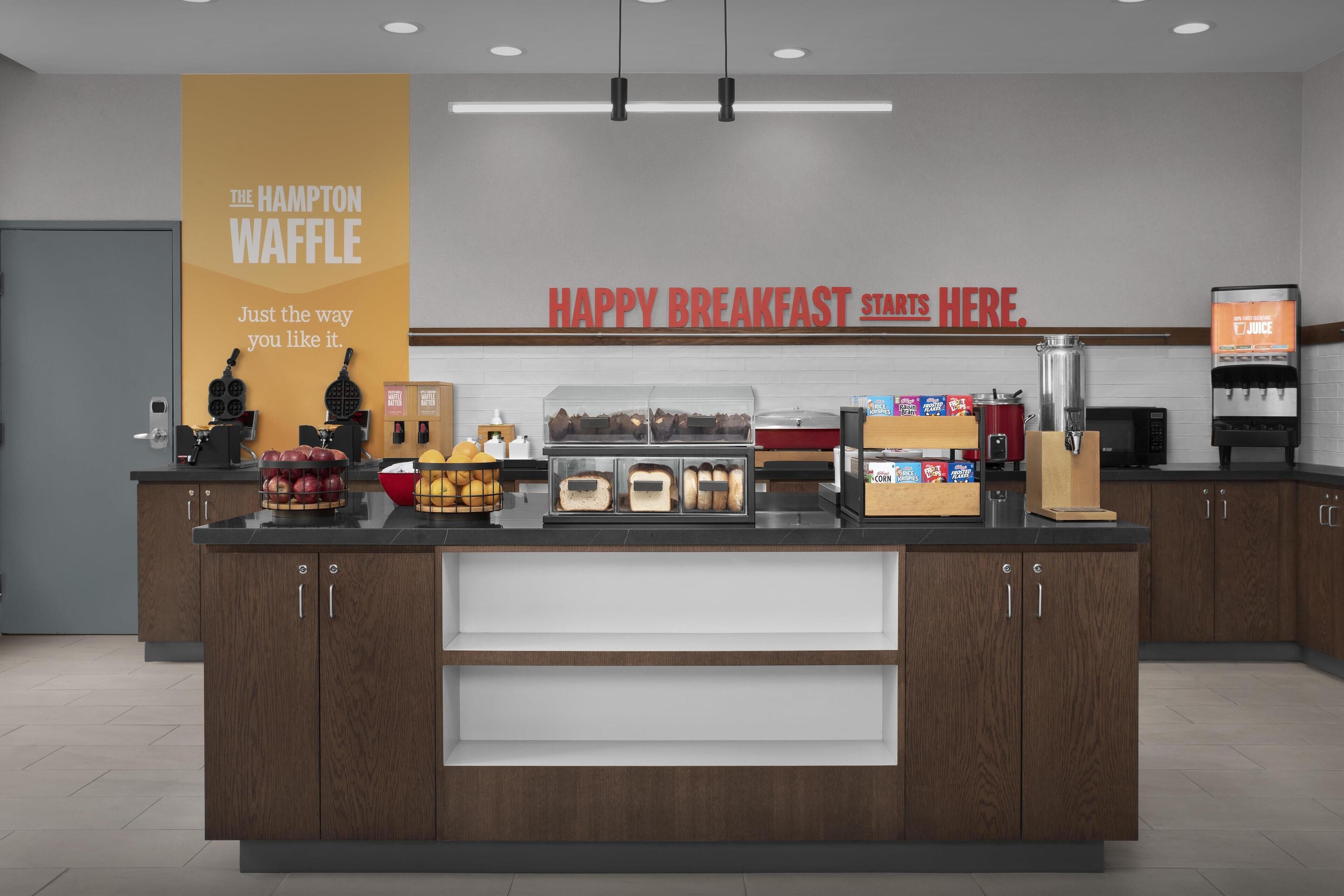 Un comptoir où l’on sert le petit-déjeuner, avec un présentoir de gaufres, un distributeur de jus et divers produits pour le petit-déjeuner.