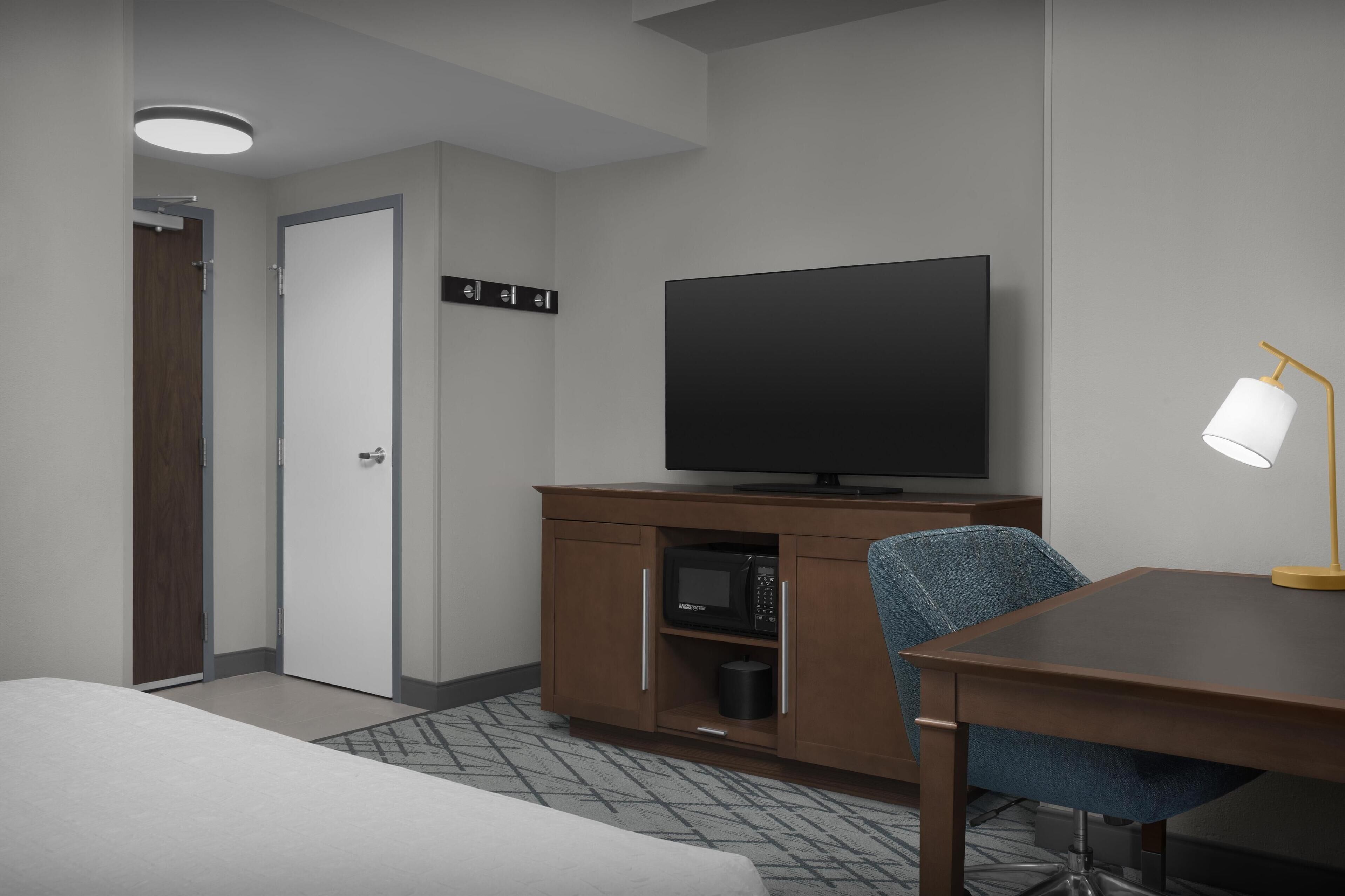 Habitación de hotel con cama, televisor sobre un mueble de madera, escritorio con lámpara y microondas.