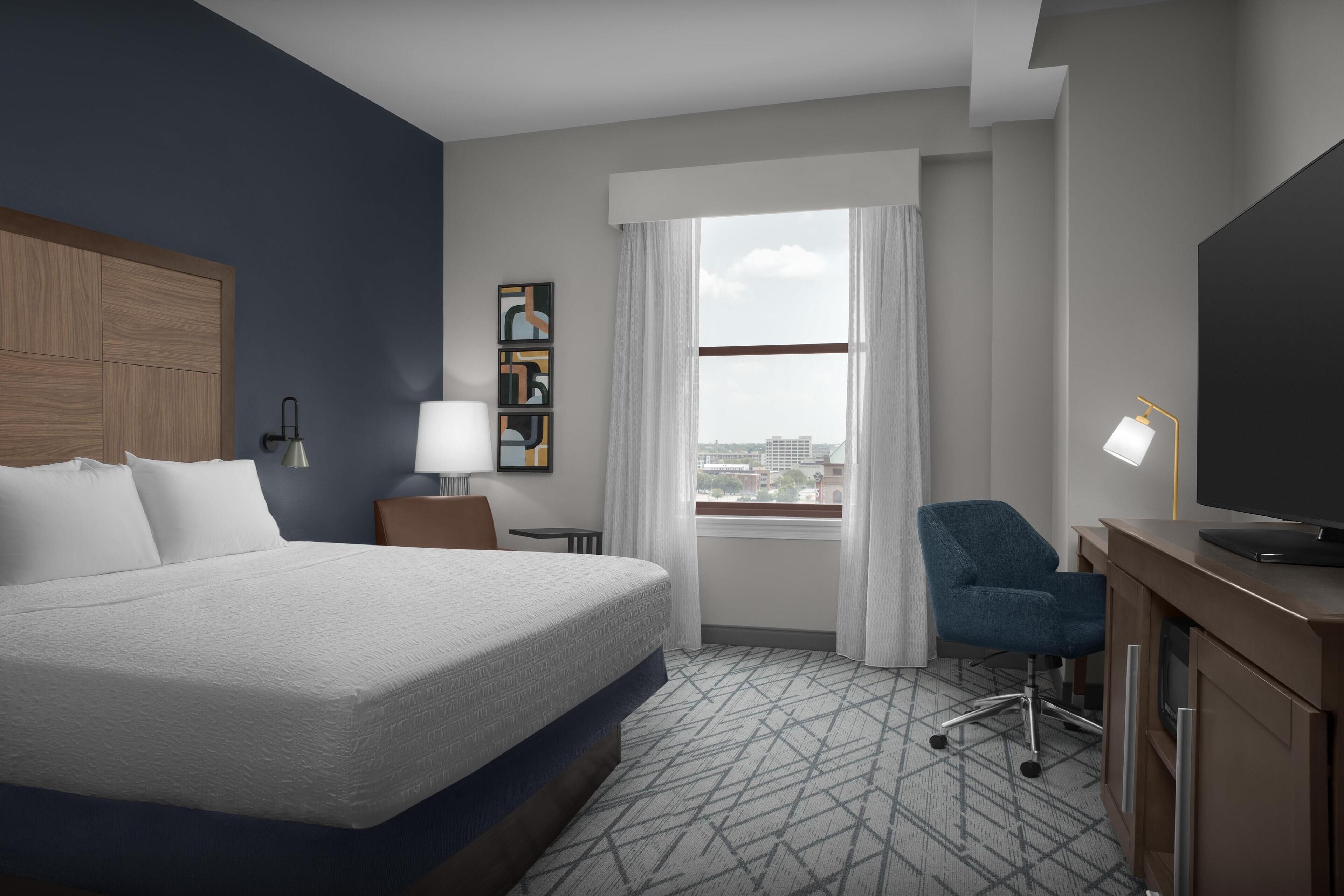 A hotel room with a large bed, a desk with a television, a chair, a window with curtains, and artwork on the wall.