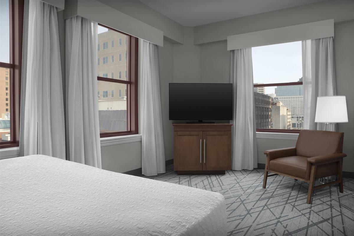 Ein Hotelzimmer mit einem großen Bett, einem Fernseher, einem Sessel und Blick auf die Stadt durch große Fenster.