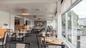 A hotel dining area with multiple tables and chairs, a view of the city through large windows, and a ceiling with patterned light fixtures. - Bay Plaza Hotel (Wellington)