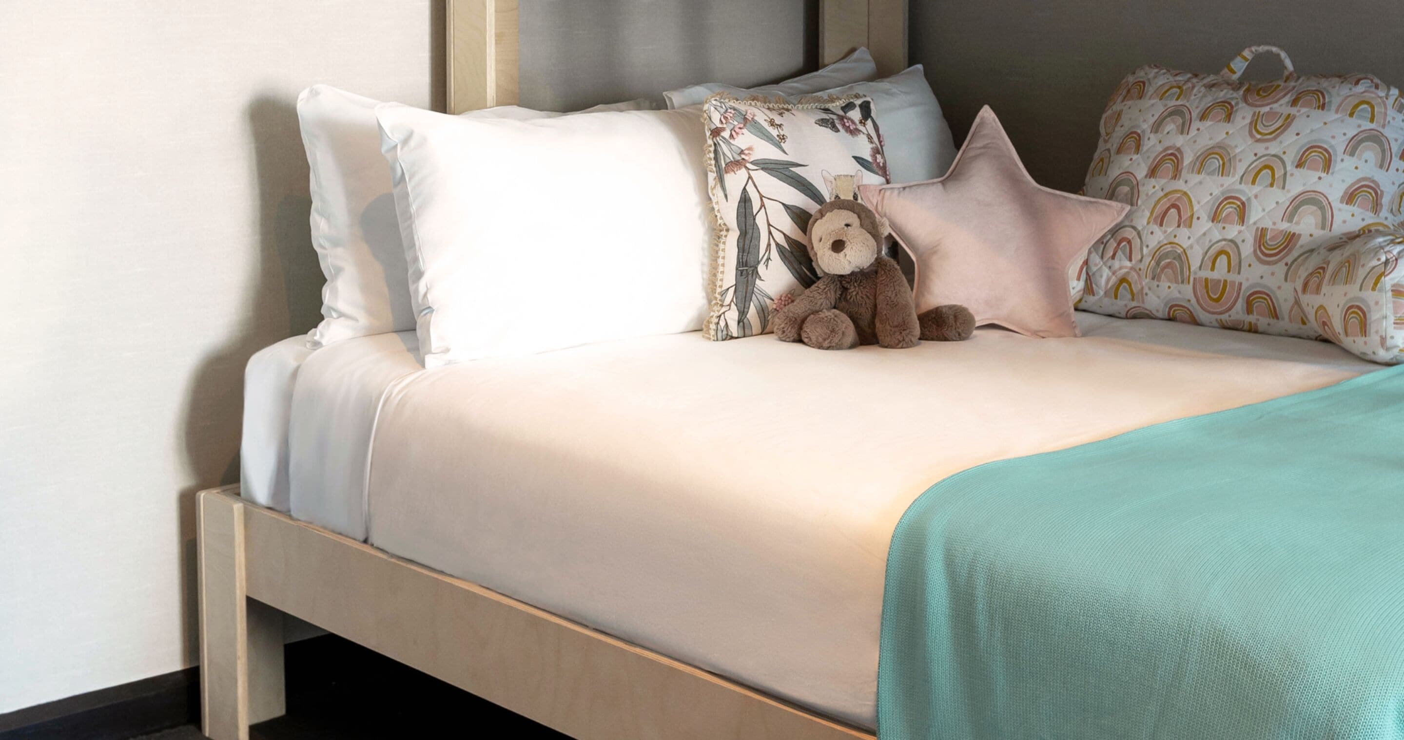 Een net opgemaakt bed met een lichtkleurige deken, een blauwgroene plaid en een teddybeer.