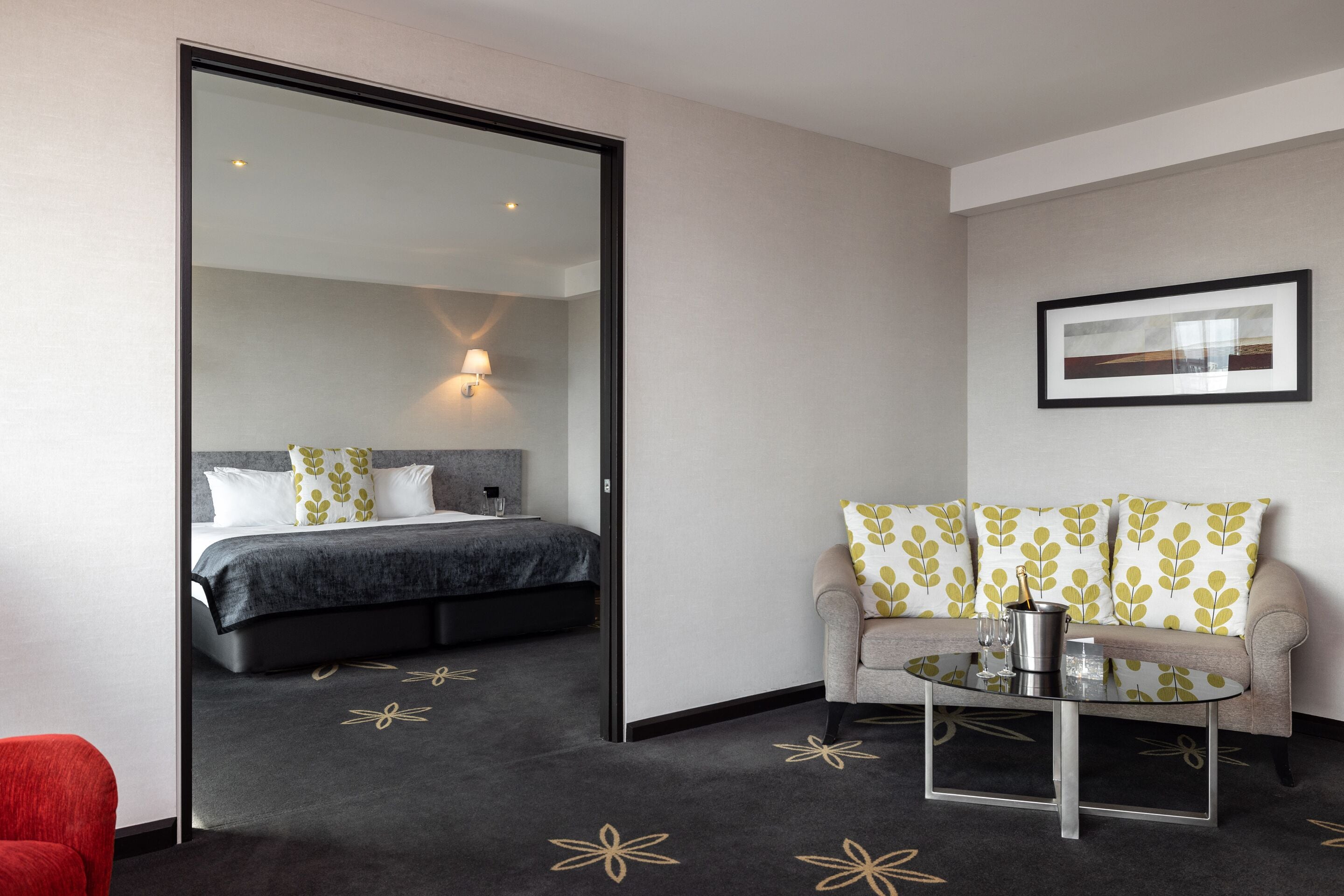 A modern hotel room with a bed, a sofa, a glass coffee table, and a red chair.