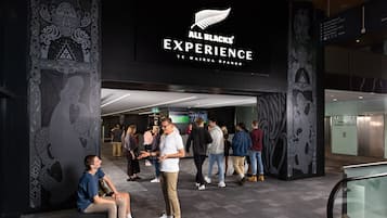 Entrada de un hotel con un cartel que dice 'All Blacks Experience' y una frase en idioma maorí. Hay gente reunida afuera, algunas sentadas y otras de pie.