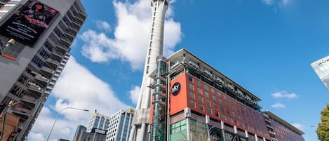 Un edificio moderno con una torre destacada, amplios ventanales y una calle concurrida llena de coches y peatones.