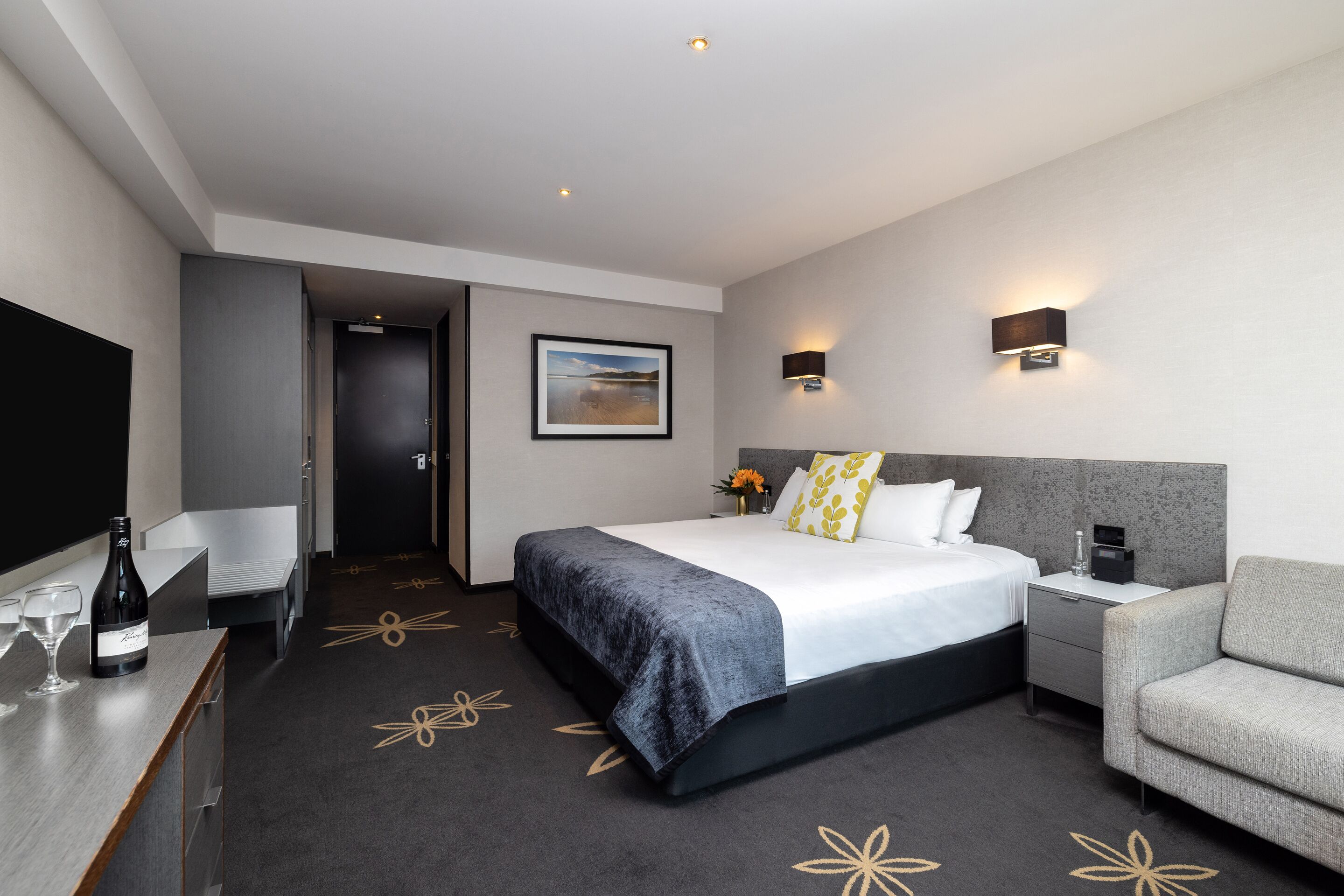 A modern hotel room with a large bed, bedside tables, a seating area, and a TV.
