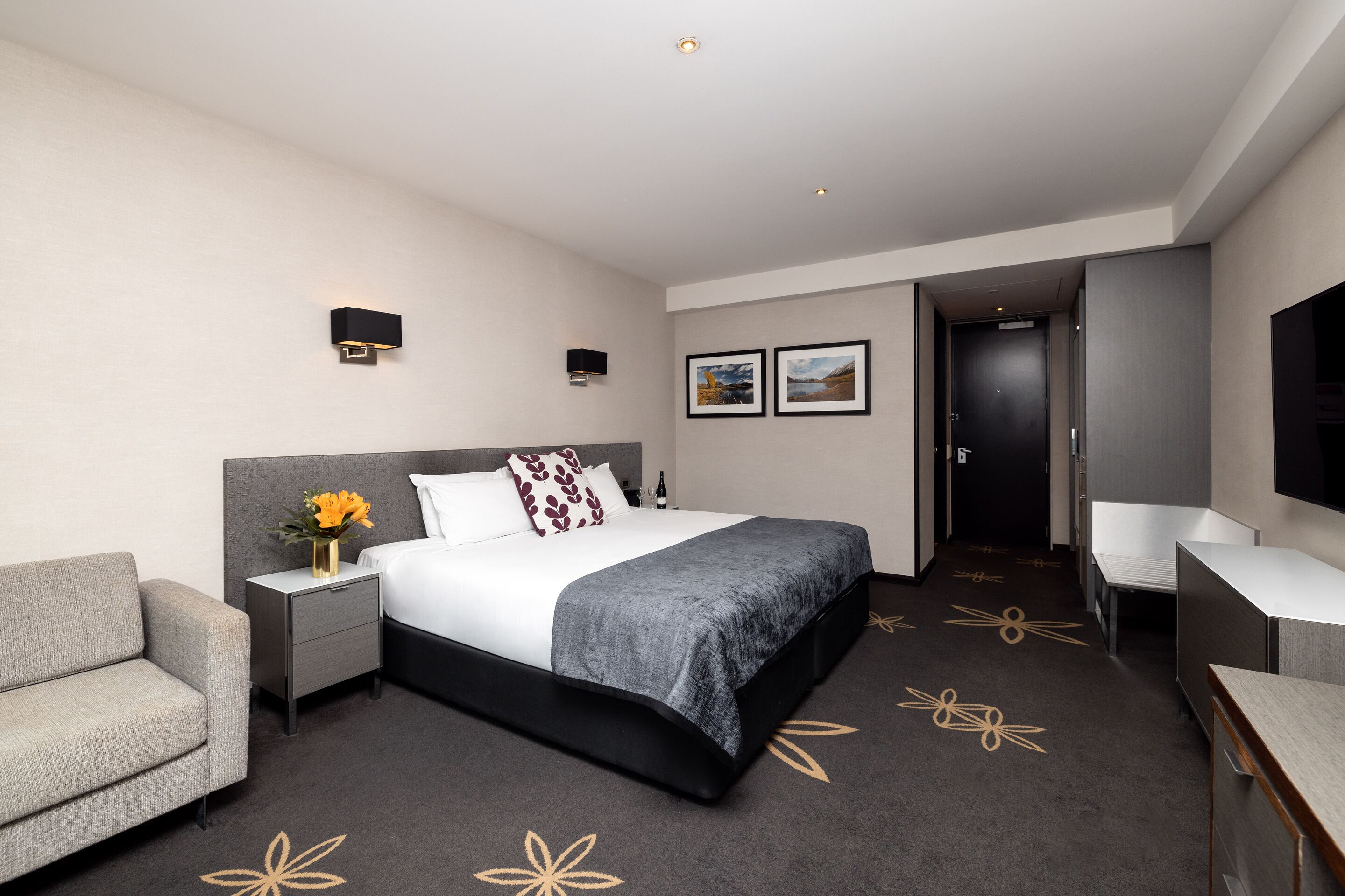 A modern hotel room with a large bed, a bedside table with a vase of flowers, a grey sofa, a desk, and a flat-screen TV.