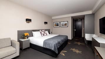 Une chambre d’hôtel moderne avec un grand lit, une table de chevet agrémentée d’un vase de fleurs, un canapé gris, un bureau et un téléviseur à écran plat.