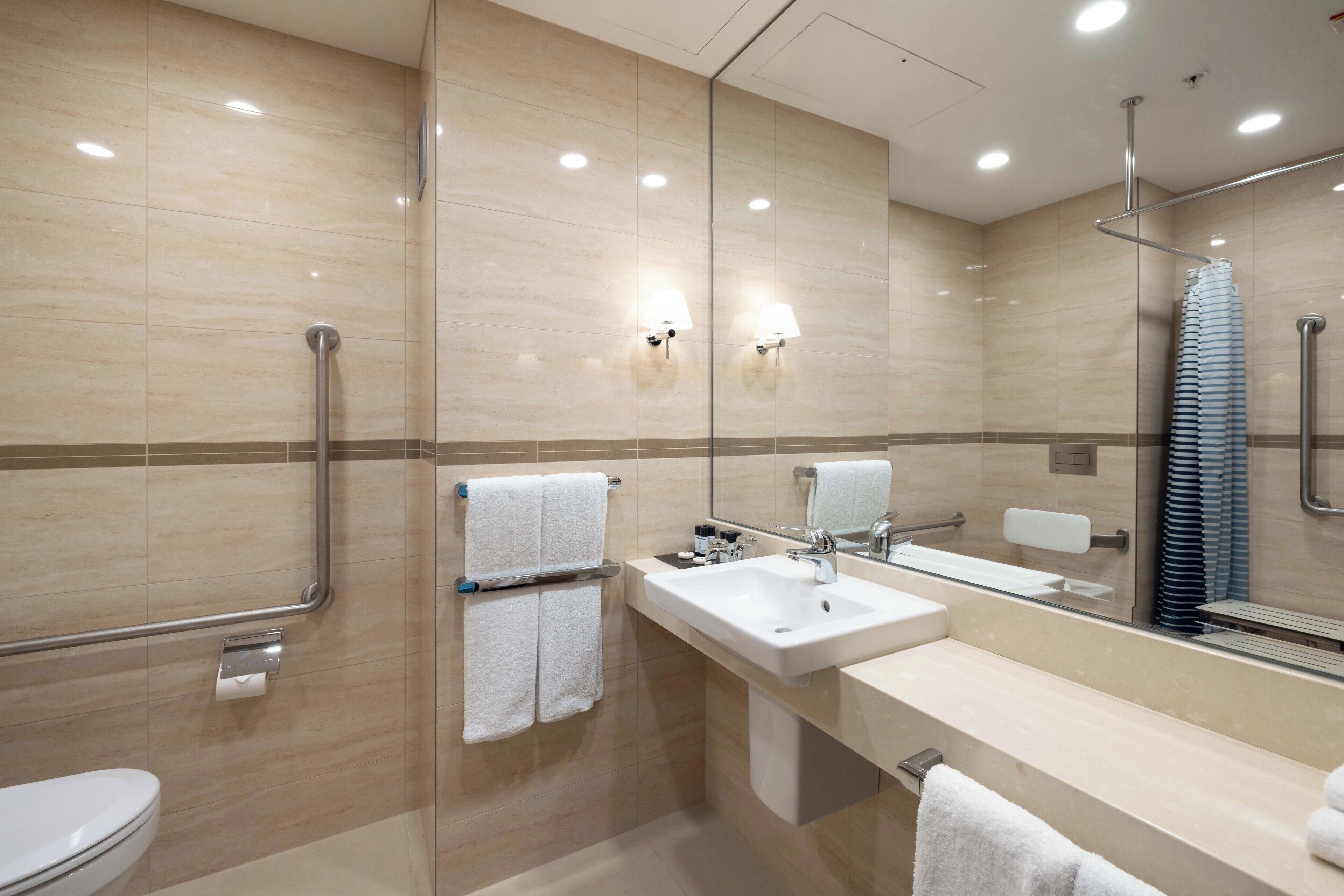 A modern bathroom with a toilet, sink, and mirror. It includes a handrail, towel rack, and a towel dispenser.