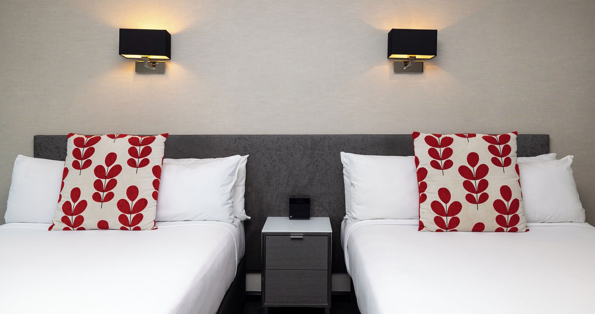 A hotel room with two beds, each with white bedding and red leaf-patterned pillows. There are two wall-mounted nightstands with black lamps.