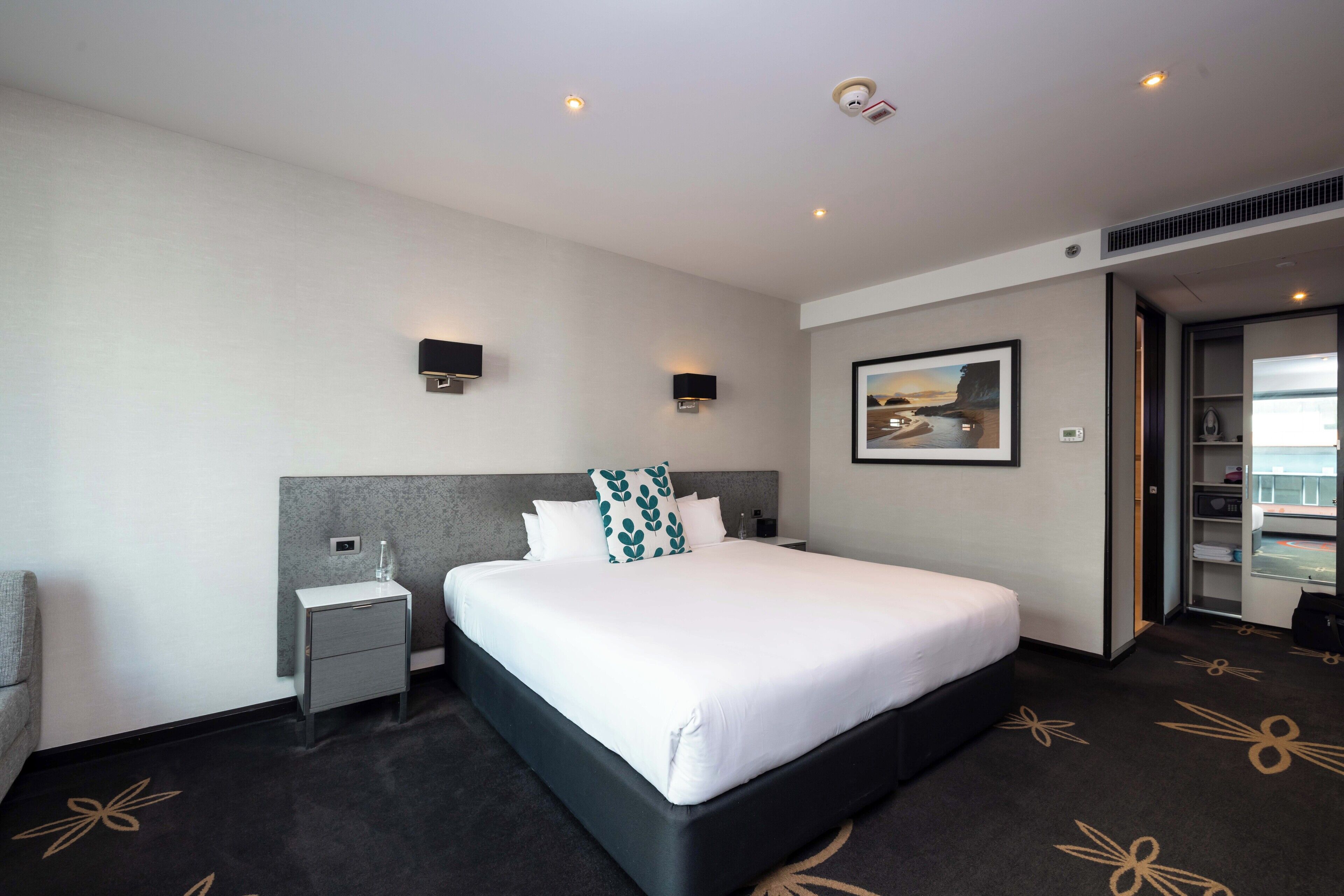A hotel room with a large bed, a nightstand, a wall-mounted lamp, and a framed picture above the bed.