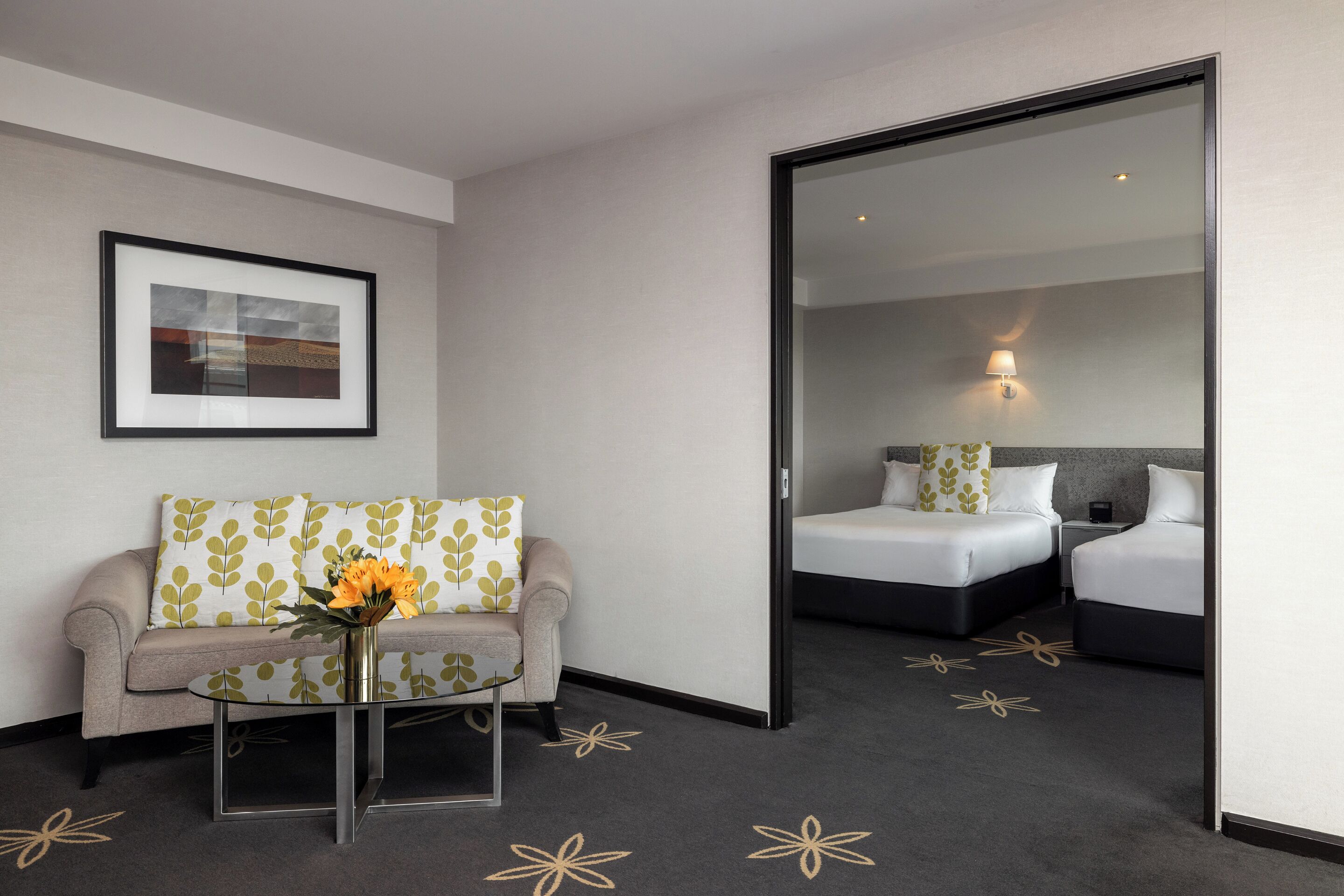 Une chambre d’hôtel avec un canapé, une petite table ornée d’un vase de fleurs et deux lits, ainsi qu’un miroir et une lampe fixée au mur.
