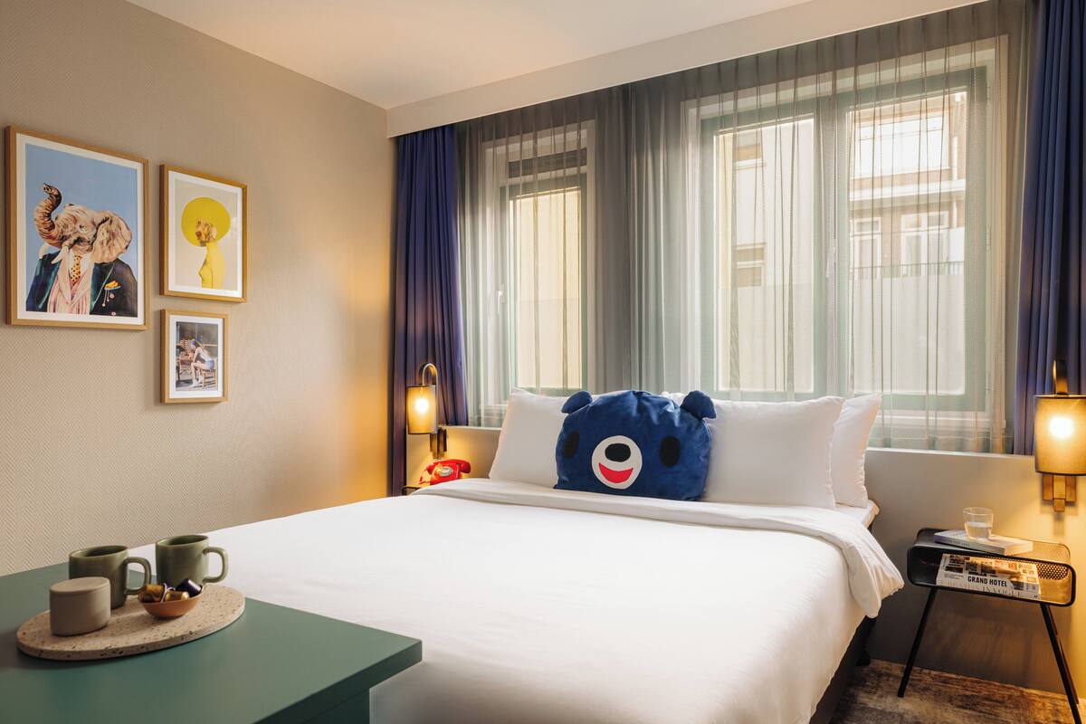 Una habitación de hotel con cama, mesita de noche con lámpara, ventana con cortinas y decoración en la pared.