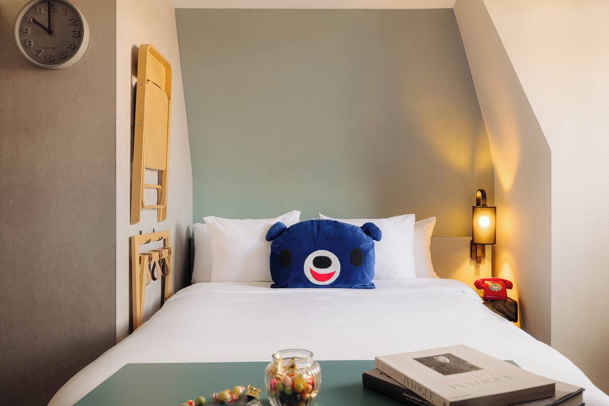 Habitación de hotel con cama, un reloj, una lámpara, un cojín con forma de osito de peluche, un frasco de vidrio y un libro.