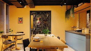 A dining area with a wooden table, chairs, a potted plant, and a kitchen area with a counter and hanging plants. - Leonardo Hotel Amsterdam Leidse Square (Amsterdam)