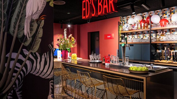 A bar with a red sign, bar stools, a counter with a vase of flowers, and shelves with bottles and glasses. - Leonardo Hotel Amsterdam Leidse Square (Amsterdam)