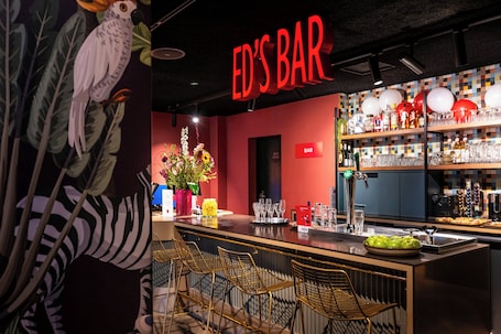 A bar with a red sign, bar stools, a counter with a vase of flowers, and shelves with bottles and glasses.