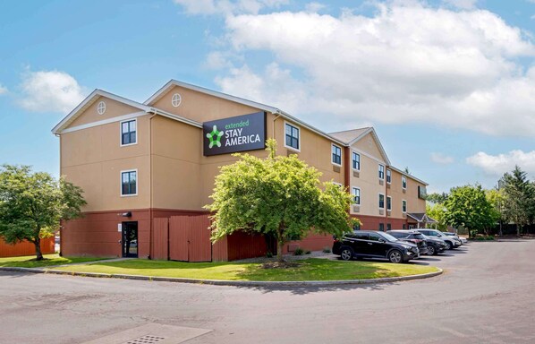A Stay America hotel with a sign, parking lot, and a tree in front. - Extended Stay America Suites Syracuse Dewitt (East Syracuse)