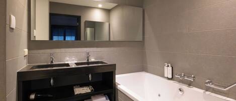 A modern bathroom with a large bathtub, a dark vanity with a mirror, and a shelf with towels.
