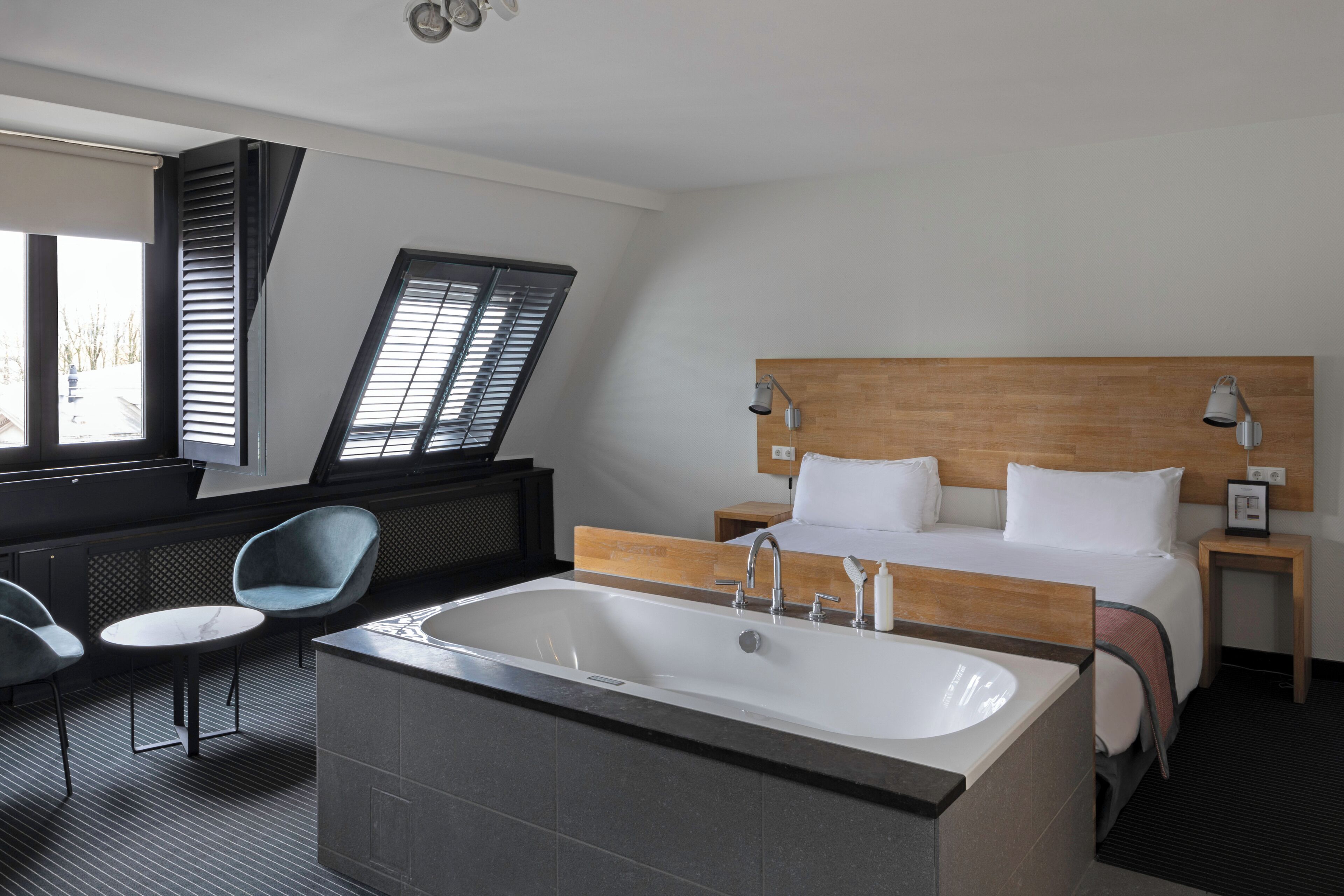 Camera d'albergo con una grande vasca da bagno, un letto con lenzuola bianche, una testiera in legno, un piccolo comodino con una lampada e una zona salotto con una poltrona e un tavolino.