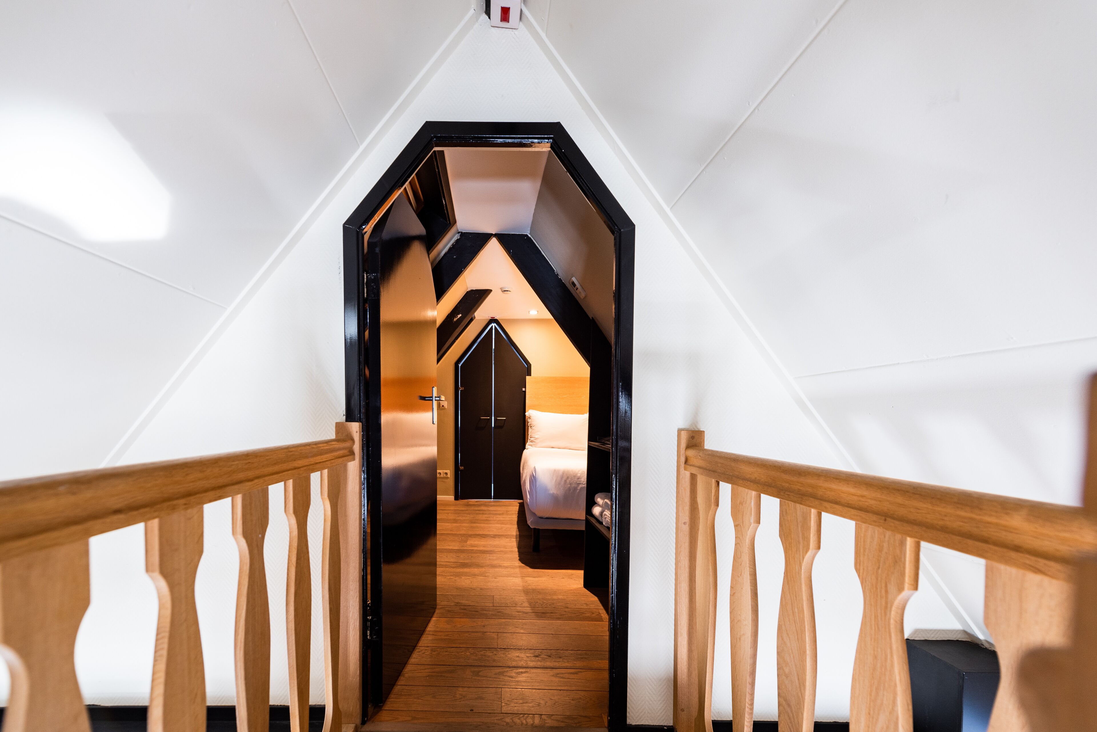 A narrow, wooden-floored hallway leading to a bedroom with a bed and a nightstand.