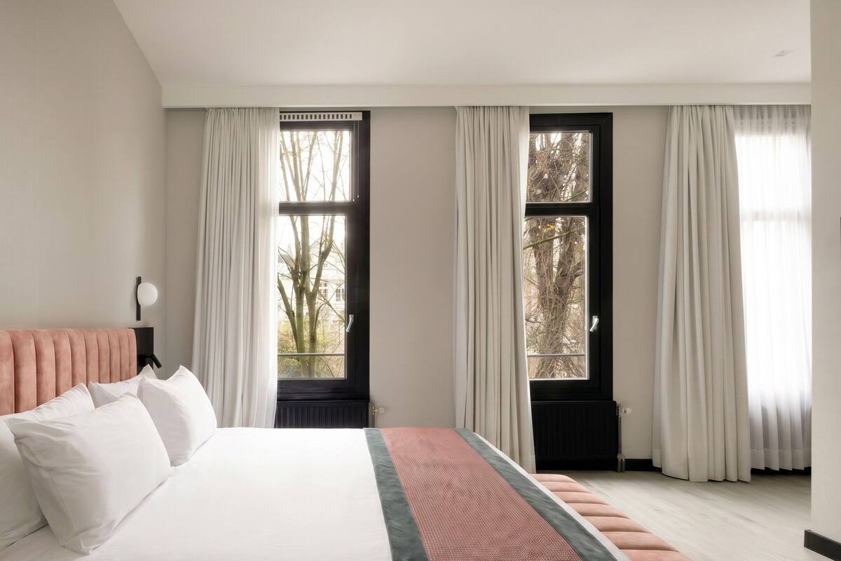 A bedroom with a bed, two windows with curtains, a radiator, and a nightstand with a lamp.