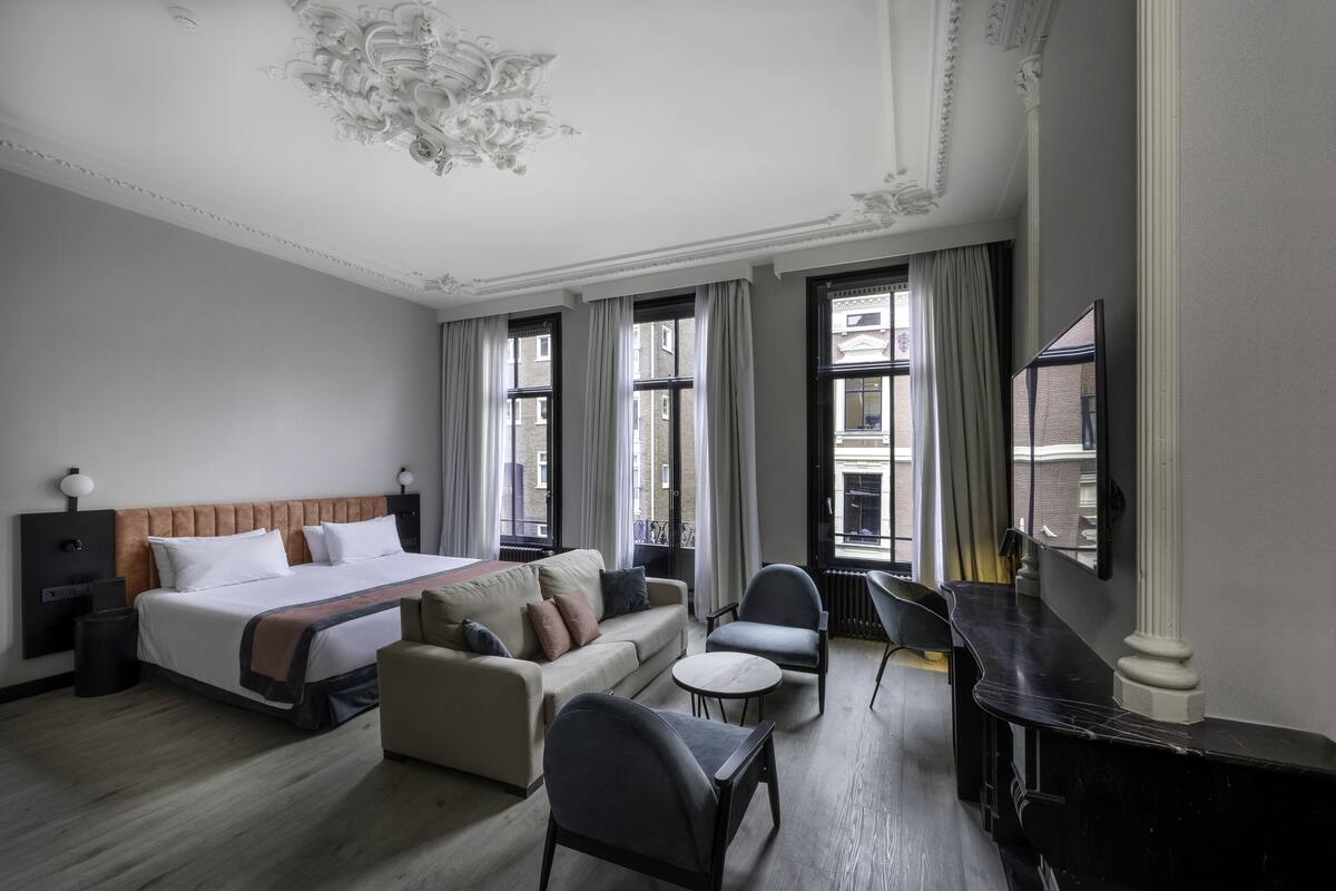 A hotel room with a bed, a sofa, a desk, and a chair. There are windows with curtains, a chandelier, and a television.