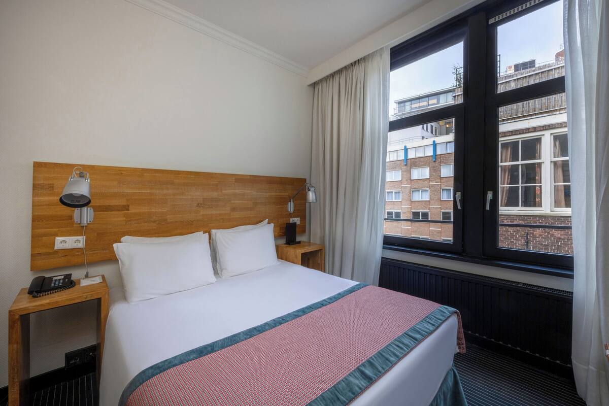 A hotel room with a wooden headboard, a large bed with white bedding, a bedside table with a lamp, and a window with a view of buildings.