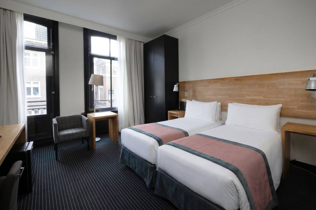 A hotel room with two beds, a desk, a chair, a nightstand, and a window with curtains.