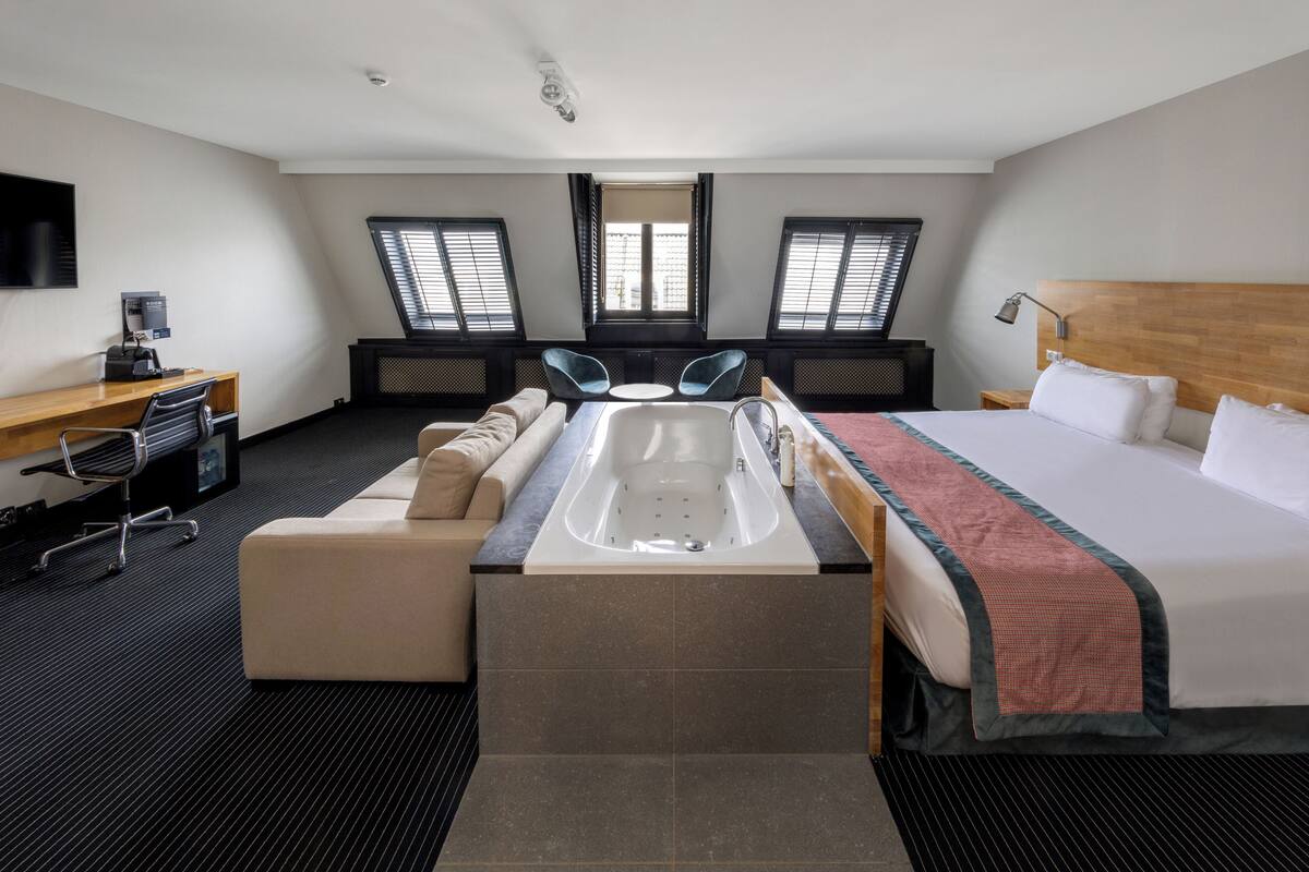 A hotel room with a bed, a desk, a chair, a bathtub, and two windows.