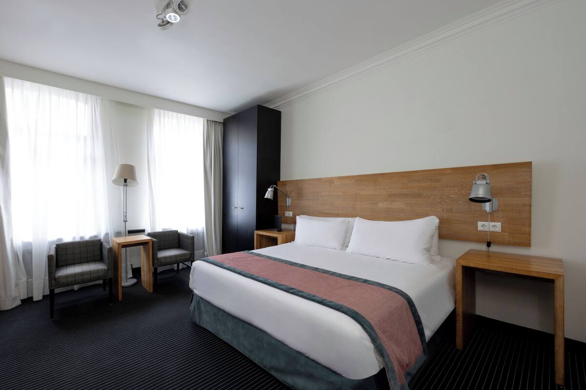A hotel room with a large bed, two chairs, a small table, a nightstand, and a wooden headboard.