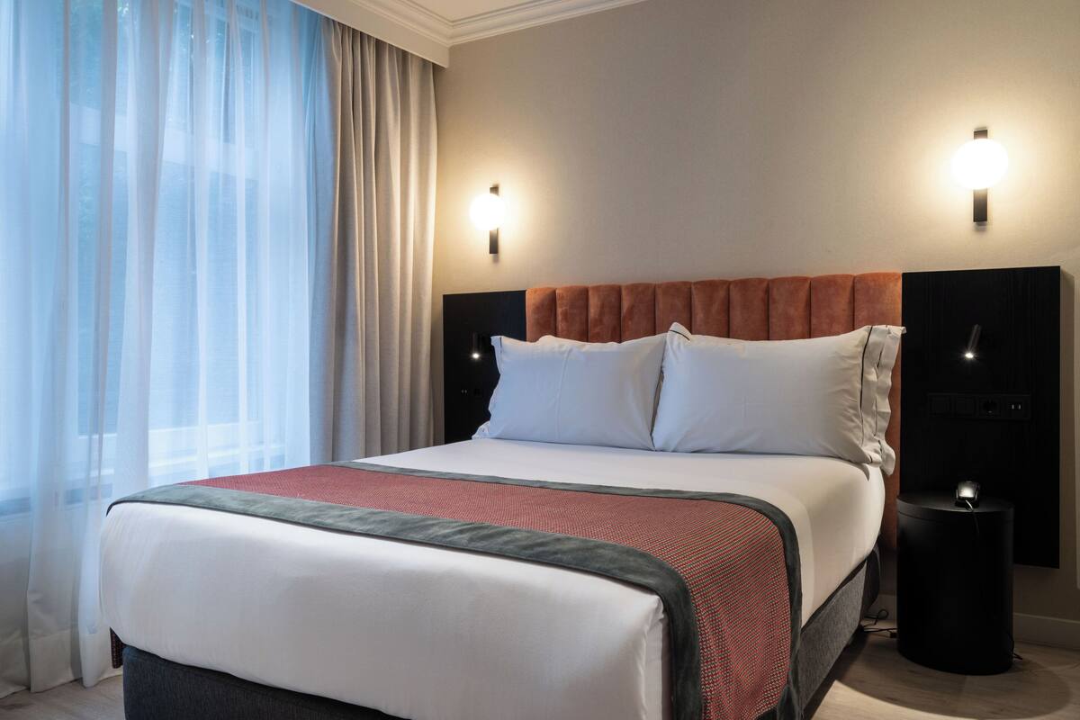 A hotel room with a large bed, two wall-mounted lights, a bedside table, and a window with sheer curtains.