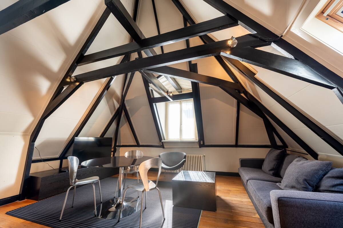 A modern loft bedroom with exposed wooden beams, a large window, a sofa, a coffee table, and a dining table with chairs.